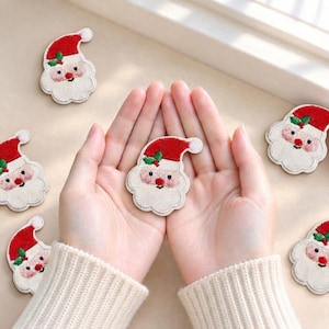 Könnte beinhalten: Mehrere gestickte Weihnachtsmann-Aufnäher mit roter Mütze samt weißem Bommel, weißem Bart und einem kleinen grünen Stechpalmenblatt. Jeder Aufnäher hat einen gewellten Rand und ein freundliches Gesicht mit roten Wangen und Nase.