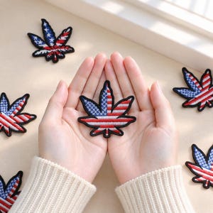 Puede incluir: Una colección de parches bordados con forma de hojas de cannabis, cada uno con un patrón de bandera estadounidense en rojo, blanco y azul con un borde negro. Los parches están dispuestos de forma dispersa, con uno centrado en el encuadre.
