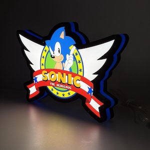 May include: A Sonic the Hedgehog LED wall sign featuring the blue character inside a green circle with white wings. A red banner across the front displays the yellow text SONIC and white text THE HEDGEHOG, accented by blue stars on a white background.