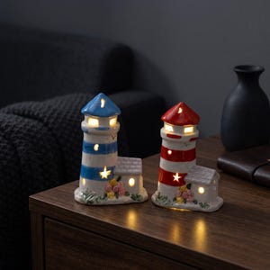 May include: Two ceramic lighthouse figurines with internal illumination. One features blue and white horizontal stripes with a blue roof, while the other has red and white stripes with a red roof. Both include small star and moon cutouts and floral accents.