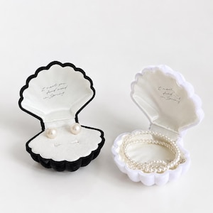 Könnte beinhalten: Zwei muschelförmige Schmuckkästchen, eines in Schwarz und eines in Weiß, mit weißem Samt ausgekleidet. Das schwarze Kästchen enthält zwei Perlenohrringe, das weiße eine Perlenkette. Beide tragen innen den Schriftzug I meet you and in Spring.
