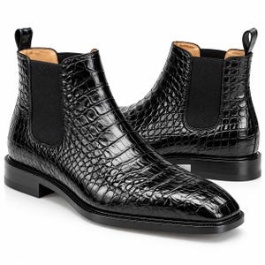 May include: A pair of black Chelsea boots featuring a glossy, embossed crocodile skin texture. The boots have black elastic side panels, a pull tab at the heel, a tan interior lining, and a solid black stacked heel with a flat, rounded toe design.