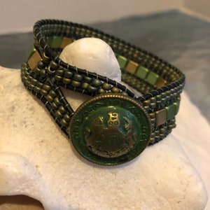 May include: Wrap bracelet featuring olive green beads, black leather, and square green accents. The bracelet is secured with a vintage green button with a crest design. The bracelet is wrapped around a white object.