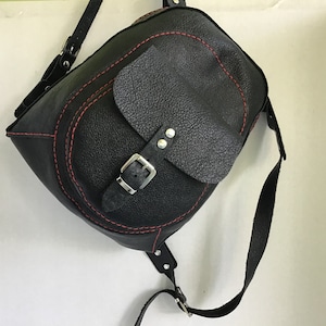 May include: Black leather backpack with a red stitch detail and a silver buckle. The bag has a flap with a smaller pocket on the front.