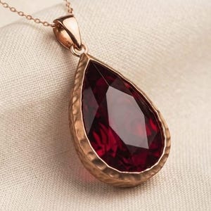 Peut inclure: Un collier pendentif en forme de goutte avec une pierre précieuse facettée rouge foncé sertie dans un cadre en métal martelé couleur or rose. Le pendentif est suspendu à une fine chaîne assortie, mettant en valeur la texture du métal.