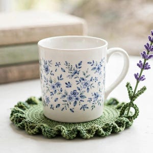 Könnte beinhalten: Ein weißer Keramikbecher mit blauem Blumenmuster steht auf einem runden, grünen Häkeluntersetzer mit gewelltem Rand. Ein kleiner, gehäkelter Lavendelzweig mit violetten Blüten und grünen Blättern ist an der Seite des Untersetzers befestigt.