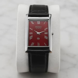 May include: A rectangular wristwatch with a deep red dial, silver-tone Roman numeral hour markers, and silver hands. The watch features a polished silver-tone case and a black leather strap with a textured finish. The dial displays Seiko Quartz text.