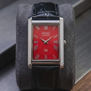 Könnte beinhalten: Eine rechteckige Seiko Quartz Armbanduhr mit einem leuchtend roten Zifferblatt mit texturiertem Guilloché-Muster, silberfarbenen römischen Ziffern als Stundenmarkierungen und silbernen Zeigern. Die Uhr hat ein poliertes silberfarbenes Gehäuse und ein schwarzes Lederarmband.
