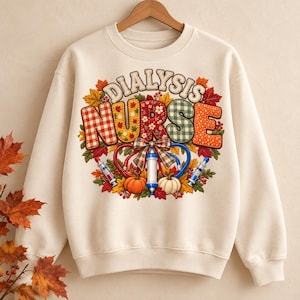 Peut inclure: Un sweat-shirt à col rond couleur crème arborant un motif avec le texte DIALYSIS NURSE en grandes lettres à motifs. Le design inclut des feuilles d'automne, des citrouilles, un nœud à carreaux et du matériel médical comme des seringues.