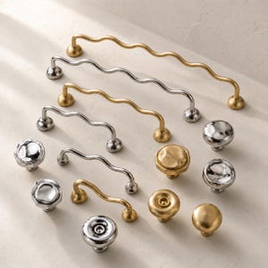 May include: A collection of cabinet hardware including wavy drawer pulls and round knobs in brushed gold and polished silver finishes. The set features various lengths of curved handles and circular knobs with smooth or textured surfaces.