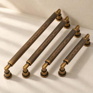 May include: Four antique brass cabinet pulls with a knurled texture and cylindrical shape. Each pull features the engraved text "Custom Engraved Pulls" along the center, flanked by small diamond-shaped decorative accents on both sides.