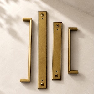 May include: Four brass-toned metal hardware pieces consisting of two U-shaped cabinet pulls and two rectangular flat plates. Each piece features a brushed metallic finish and visible screw heads for mounting, arranged vertically on a light surface.