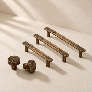 May include: Three bronze-toned metal drawer pulls of varying lengths and two round, fluted knobs. Each pull features a textured surface, a horizontal line, and the engraved text "Custom Engraved Pulls" with diamond-shaped accents.