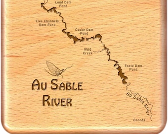 May include: A wooden map of the Au Sable River in Michigan, showing the river's course and the locations of various dams and ponds. The map is engraved with the text "Au Sable River" and a stylized image of a mayfly.