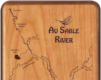 May include: A wooden plaque with a map of the Au Sable River in Michigan. The map shows the river's course, including its tributaries, and features various points of interest, such as bridges and a lake. The plaque also includes the text "Au Sable River" and a silhouette of a mayfly.