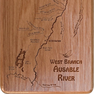 May include: A wooden plaque with an engraved map of the West Branch Ausable River in New York. The map shows the river's tributaries, including Little BlackBrook, Brown Brook, Pettigrew Brook, Bridge Brook, Stephenson Brook, Red Brook, White Brook, Beaver Brook, and East Branch Ausable River. The map also shows the location of Whiteface Mountain, Wilmington, High Falls Gorge, Lake Placid, and Outlet Brook. The plaque also features the text "West Branch Ausable River" and a stylised image of a mayfly.