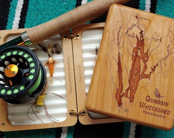 May include: A wooden fly fishing box with a map of the Quabbin Watershed in Massachusetts on the lid. The box is open and contains a fly fishing rod, reel, and flies. The text "Handcrafted in Montana" is visible on the side of the box.
