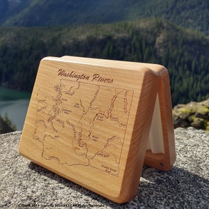 May include: A wooden fly fishing box with a map of Washington state rivers engraved on the lid. The box is open and the lid is propped up against a rocky surface. The background is a scenic view of mountains and trees.