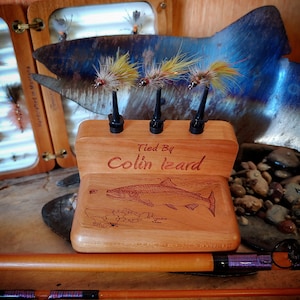 May include: A wooden fly fishing fly tying display stand with three flies mounted on it. The stand is engraved with the text "Tied By Colin Izard".