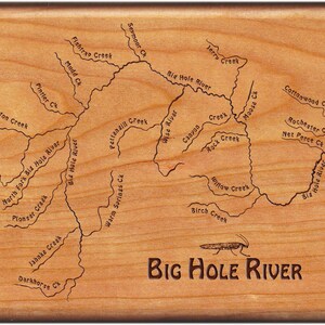 May include: A wooden map of the Big Hole River in Montana, USA. The map shows the river and its tributaries, including the names of the creeks and rivers. The map is engraved on a light brown wooden surface. The text "BIG HOLE RIVER" is engraved at the bottom of the map.