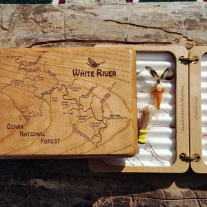 May include: A wooden fly fishing box with a map of the White River in the Ozark National Forest on the lid. The box is open and contains a variety of flies. The box is handcrafted in Montana.