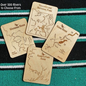 May include: Four wooden plaques with engraved maps of different rivers. The plaques are on a green and black striped surface. The text on the plaques reads "Yellowstone National Park", "Kenai River", "Watauga River", and "Henry's Fork River".