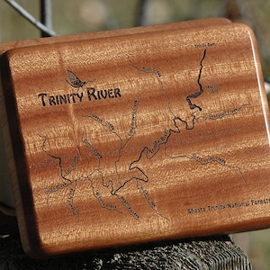 May include: A wooden box with a detailed map of the Trinity River and its tributaries, including Lewiston Lake, etched into the surface. The text "Trinity River" is prominently displayed, along with "Shasta Trinity National Forests". The box is made of reddish-brown wood.