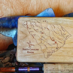 May include: A wooden box with a laser-engraved map of South Carolina rivers. The box is light brown with a natural wood grain. The map shows the major rivers and lakes in the state. The text "South Carolina Rivers" is engraved below the map.