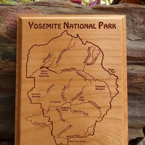 May include: A wooden plaque with a laser-engraved map of Yosemite National Park. The map shows the major rivers, creeks, and landmarks within the park. The plaque is a light brown color with a dark brown border.
