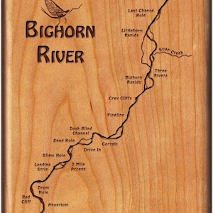 May include: A wooden plaque with a map of the Bighorn River, showing fishing spots and access points. The map is engraved with the names of the locations, including "Bighorn Access", "Last Chance Hole", "Littlehorn Rapids", "Soap Creek", "Three Rivers", "Grey Cliffs", "Pipeline", "Duck Blind Channel", "Snag Hole", "Corrals", "Drive In", "Slime Hole", "Landing Strip", "3 Mile Access", "Drum Hole", "Red Cliff", and "Aquarium". The plaque also features the text "BIGHORN RIVER" and a silhouette of a mayfly.