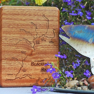 May include: A wooden box with a map of the Blackfoot River engraved on the top. The map includes the names of various creeks and rivers, including the North Fork Blackfoot River, Moisture Creek, Clearwater River, and Gold Creek. The box is sitting on a table with a metal fish sculpture and purple flowers in the background.