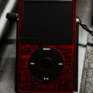 May include: A portable digital music player with a translucent red casing revealing internal circuitry. It features a black circular click wheel with white playback icons and the word MENU, a black screen, and a black headphone cable connector.