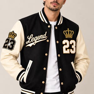 Pode incluir: Um casaco varsity preto com mangas cor de creme, apresentando o texto Legend, o número 23 e um emblema de coroa dourada no peito e na manga. O casaco tem gola, punhos e bainha canelados com riscas brancas e douradas, e botões de pressão dourados.