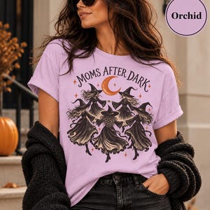 May include: A light purple t-shirt featuring a graphic print of four witches in black dresses and hats dancing in a circle under an orange crescent moon and stars, with the text "MOMS AFTER DARK" arched above the illustration in black lettering.