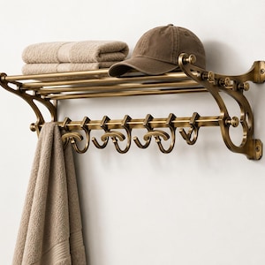May include: A wall-mounted brass towel rack with a top shelf and a row of six curved hooks. A folded beige towel and a brown baseball cap rest on the top shelf, while a second beige towel hangs from one of the hooks on the lower rail.