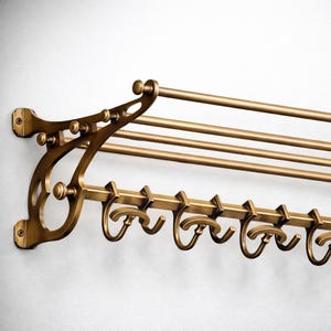May include: A wall-mounted coat rack with a brass finish, featuring a top shelf with three horizontal bars and a lower section equipped with multiple curved hooks. The structure is supported by two ornate side brackets with screw holes.