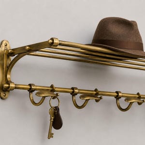 May include: A wall-mounted brass coat rack with a top shelf and multiple hooks. A brown felt fedora hat rests on the top shelf, and a single brass key with a brown leather tag hangs from one of the hooks on the lower rail of the metal rack.