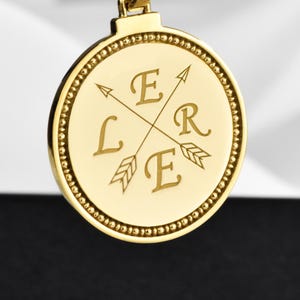 May include: A gold-toned circular pendant necklace featuring a rope chain. The pendant displays two crossed arrows and the letters E, L, R, and E engraved in the four quadrants. The edge of the circular pendant has a beaded texture detail.