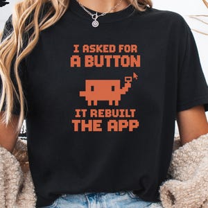 May include: A black short-sleeve t-shirt featuring orange pixelated text that reads I ASKED FOR A BUTTON IT REBUILT THE APP, accompanied by a small pixelated graphic of a creature holding a cursor icon, centered on the chest area.