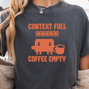 May include: A charcoal gray short-sleeve t-shirt featuring orange pixelated graphics and text. The design includes a loading bar, a blocky character, a coffee mug, and the text CONTEXT FULL COFFEE EMPTY printed in a retro digital font style.