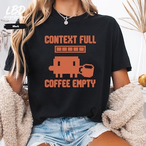 May include: A black short-sleeve t-shirt featuring an orange pixelated graphic of a small creature holding a mug. Above the graphic is the text CONTEXT FULL and below is the text COFFEE EMPTY, with a loading bar icon between the two lines.