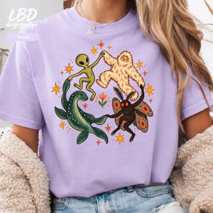 May include: A light purple t-shirt featuring a graphic print of a green alien, a beige yeti, a green sea creature, and a black moth dancing in a circle surrounded by orange stars and a small flower. The shirt has a crew neckline.