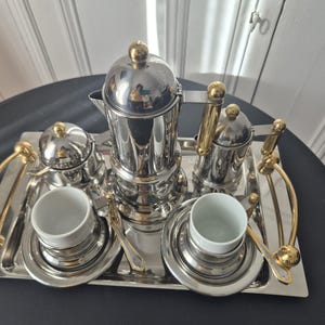 May include: A silver-toned coffee service set featuring a tall coffee pot, a lidded sugar bowl, a creamer, and two white ceramic cups with matching silver saucers. The set is arranged on a rectangular silver tray with curved gold-toned handles.