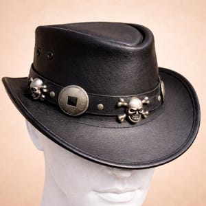 Op de afbeelding: Een zwarte leren fedora hoed met een brede band versierd met zilverkleurige metalen schedel-emblemen, gekruiste botten en een rond, muntachtig middelpunt. De hoed heeft twee kleine zwarte ventilatiegaatjes aan de zijkant van de bol.