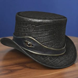 May include: A black top hat featuring a textured crocodile skin pattern across the crown and brim. A smooth black band encircles the base, accented with a single, realistic brown eye detail centred on the front, framed by a metallic gold trim.