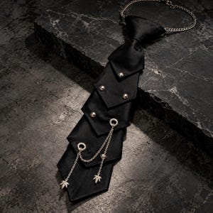 Puede incluir: Una corbata de satén negro con un diseño en capas y puntiagudo, adornada con tachuelas y ojales de metal plateado. Una fina cadena plateada cuelga de los ojales inferiores, terminando en dos pequeños dijes detallados en forma de hoja con cadena.
