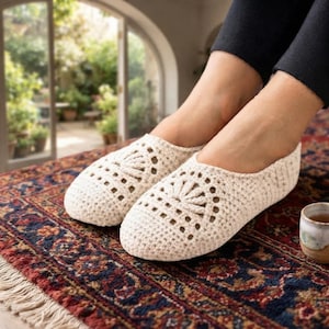 May include: A pair of off-white crochet slippers featuring a decorative fan-shaped pattern on the top of the foot. The slippers are crafted with a dense, textured stitch and have a rounded toe design, providing full coverage for the feet.