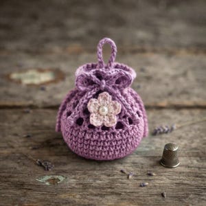 Puede incluir: Una pequeña bolsa redonda de ganchillo color lavanda con cierre de cordón y un lazo decorativo. La parte frontal está adornada con una flor de ganchillo rosa claro centrada con una perla blanca. Un dedal de metal se encuentra al lado.