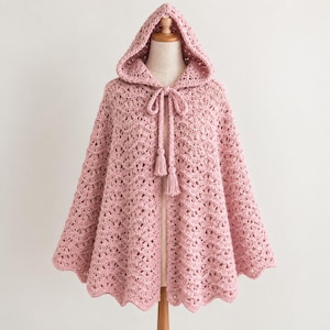 May include: A light pink crochet hooded cape featuring an intricate openwork wave pattern throughout the fabric. The garment includes a matching drawstring tie at the neckline finished with two decorative tassels hanging down the front center.