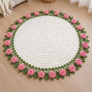 May include: A round, white crocheted rug featuring a decorative border of green crocheted leaves and three-dimensional pink rose blossoms arranged in a circular pattern around the edge of the white center section of the textile floor covering.